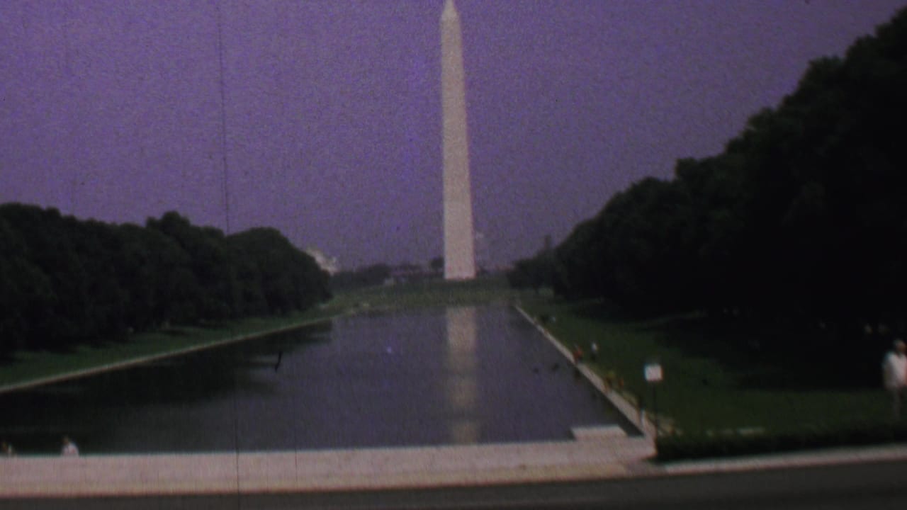 1969 8mm view of the Washington Monument reflected in the Lincoln Memorial Reflecting Pool, framed by dark trees, green lawns, and a purple-tinged sky.