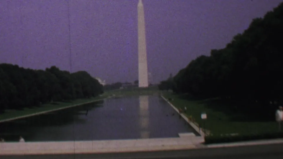 1969 8mm view of the Washington Monument reflected in the Lincoln Memorial Reflecting Pool, framed by dark trees, green lawns, and a purple-tinged sky.