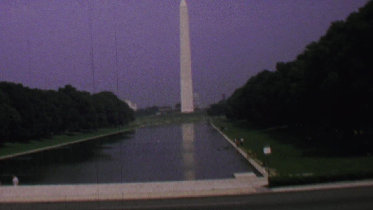 1969 Washington, DC home-movie frame showing the Washington Monument above the Reflecting Pool with trees, lawns, a small sign, and a distant visitor on the path.