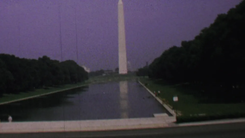 1969 Washington, DC home-movie frame showing the Washington Monument above the Reflecting Pool with trees, lawns, a small sign, and a distant visitor on the path.