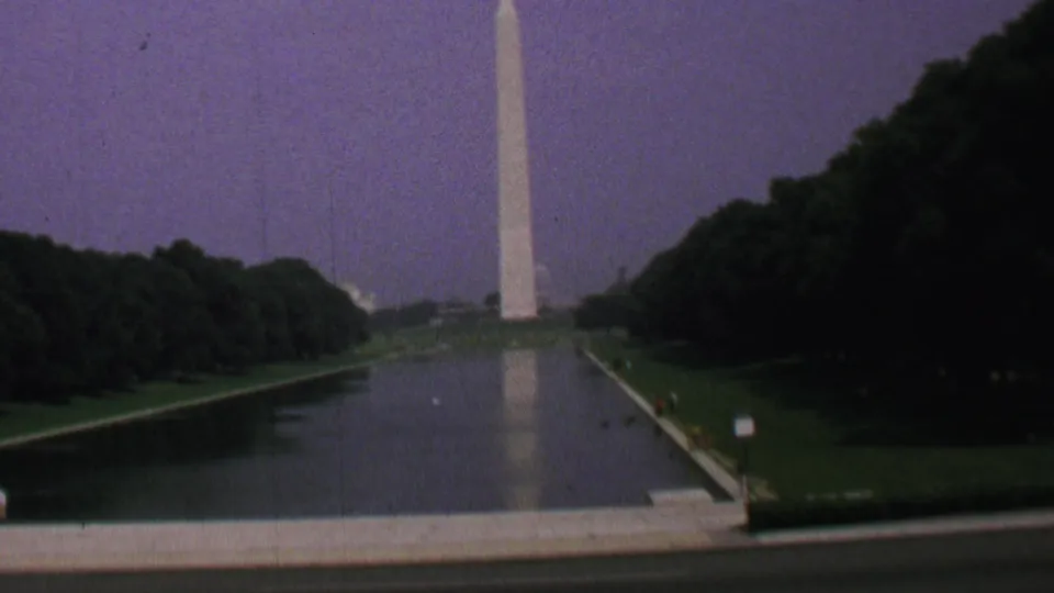 Soft 8mm frame of the Washington Monument and its reflection across the Lincoln Memorial Reflecting Pool, bordered by trees, lawns, a foreground walkway, and small signs.