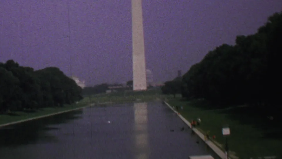 1969 8mm home-movie view of the Washington Monument and Reflecting Pool with tree-lined lawns, small posted signs, and tiny distant visitors near the walkway.