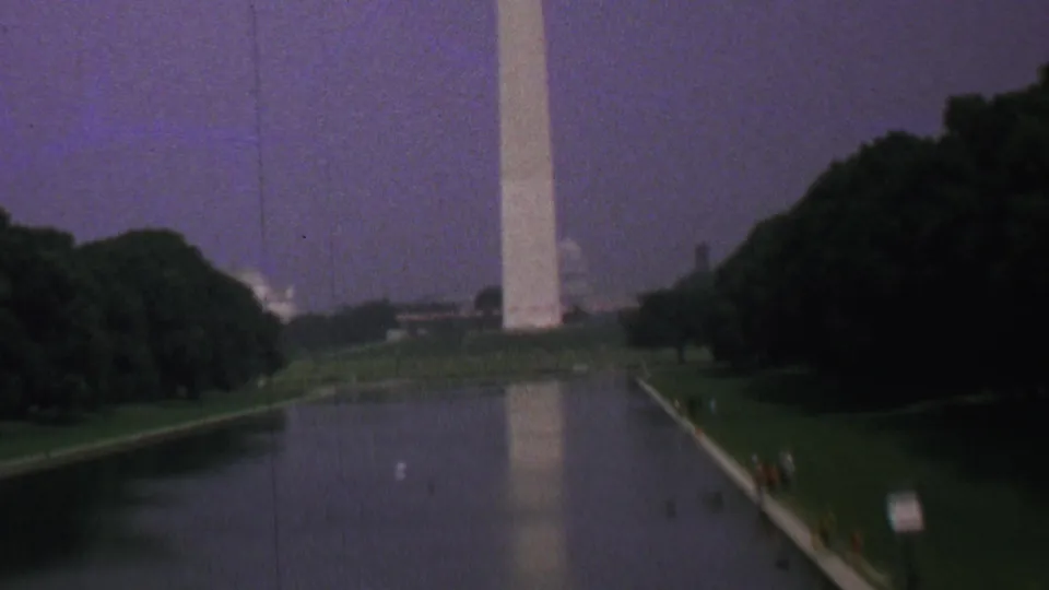 1969 8mm home-movie frame of the Washington Monument seen across the Reflecting Pool in Washington, DC, with tree-lined lawns and small visitors along the edge.