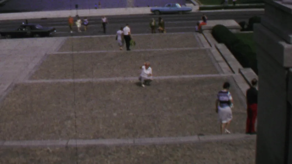 Wide visitor-shot frame of the Lincoln Memorial plaza in Washington, DC, with scattered tourists, a roadway, and 1960s-era cars visible below.