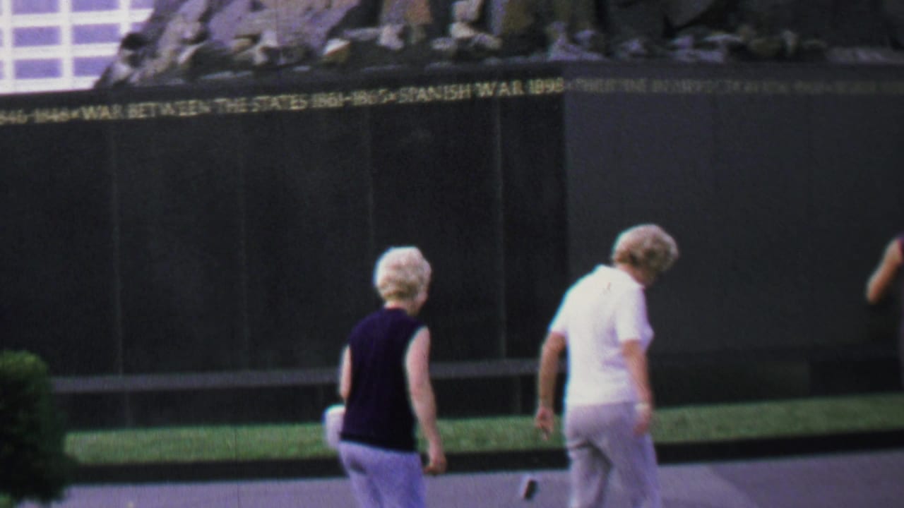 Two older women walk past a dark inscribed memorial wall with the Reflecting Pool area nearby.