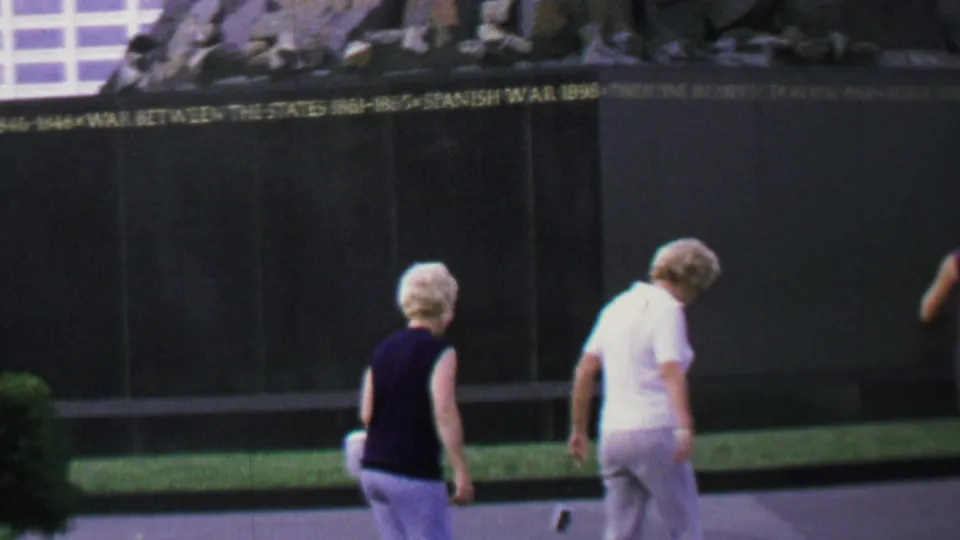 Two older women in light pants walk along a paved path beside an engraved memorial wall in a Washington, D.C. landmark setting.