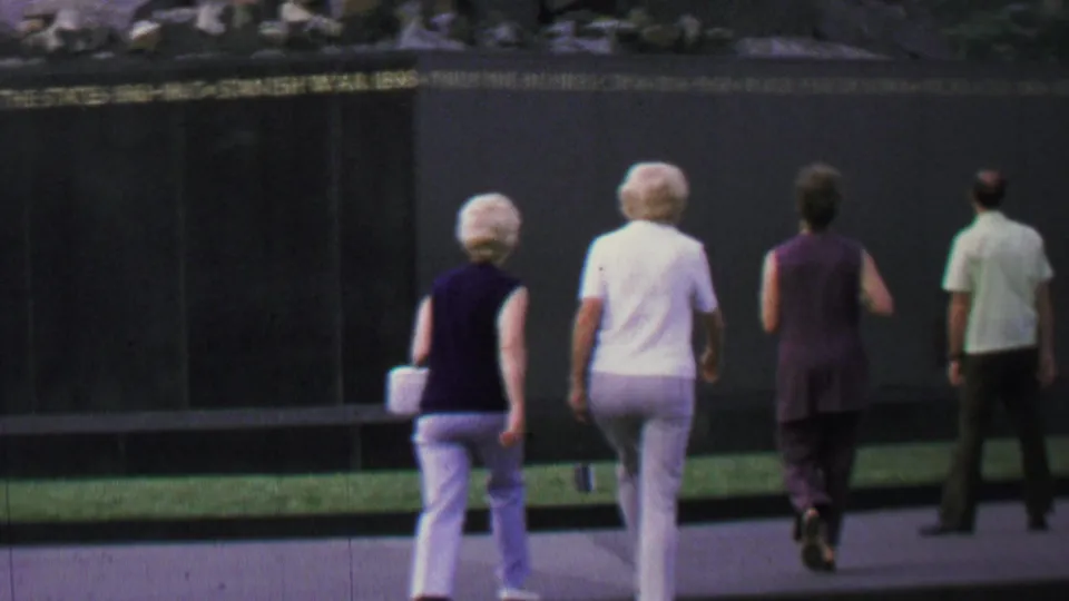 A small group of older visitors and adults walk toward an engraved memorial wall during a 1960s Washington, D.C. sightseeing stop.