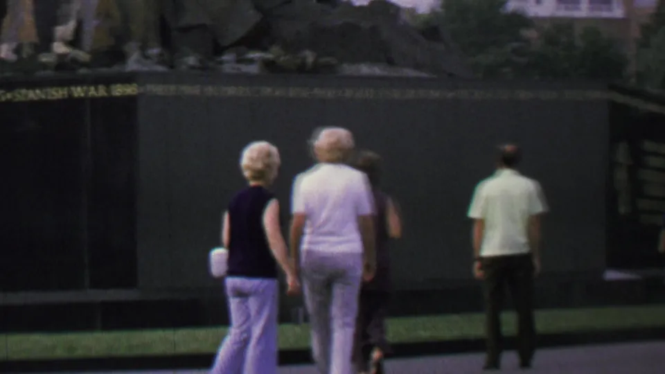 Visitors in casual late-1960s clothing stand and walk beside a dark inscribed memorial wall in a public Washington, D.C. monument area.