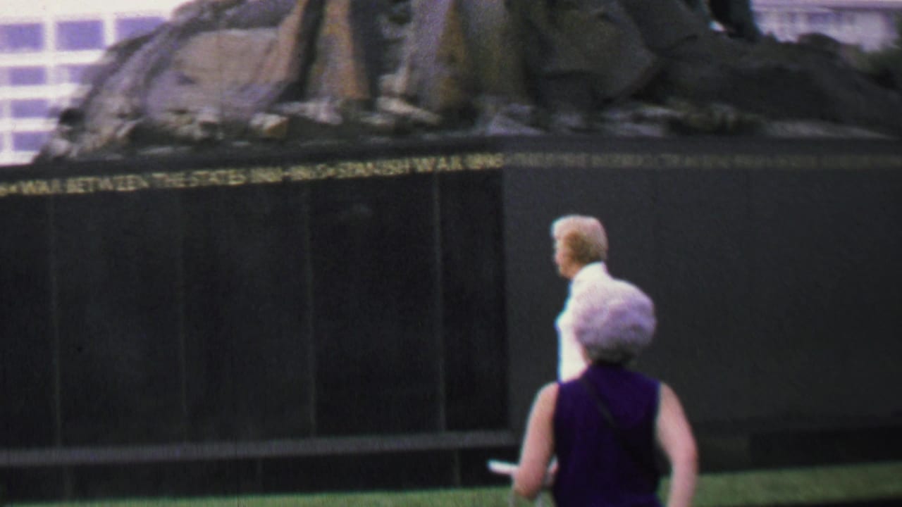 In supported 1969 Washington, DC home-movie footage, older women pass a dark public war memorial with gold inscriptions and a sculptural upper section.