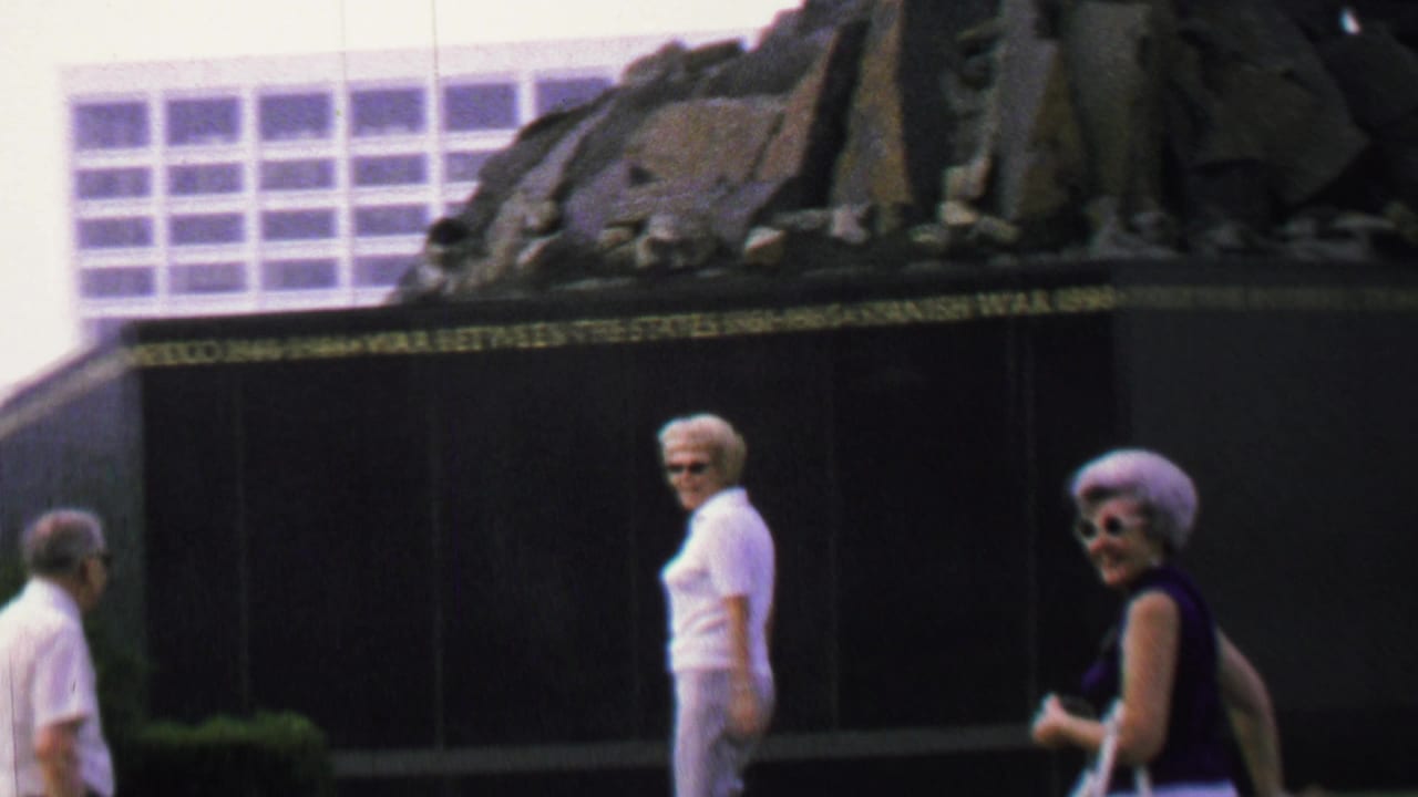 A woman in sunglasses looks toward the camera while another woman walks beside the memorial wall.