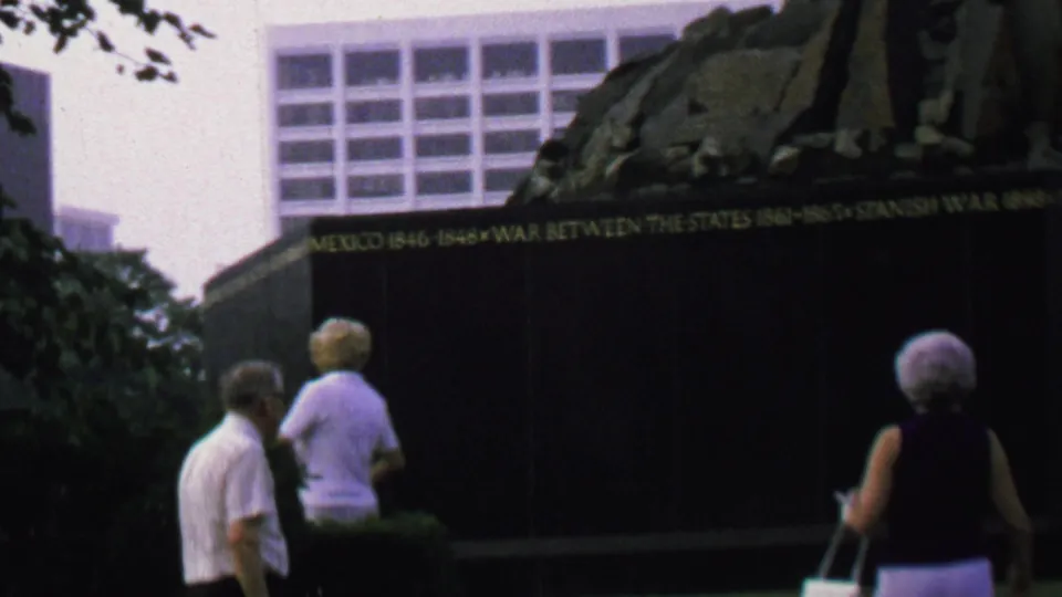 Several older visitors move across the foreground near shrubs and grass as the memorial wall shows legible gold references to conflicts such as the Mexican War and Spanish-American War.