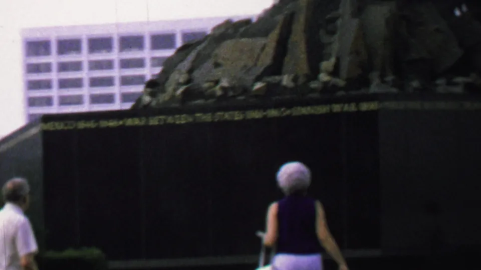 A white-haired woman in a sleeveless dark top walks away toward the dark memorial wall, with gold lettering, grass, and a modern building behind the monument.