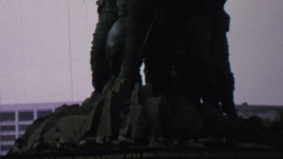 The camera frames the lower and central figures of the Iwo Jima Memorial in silhouette, with the memorial base and a background building visible behind the sculpture.