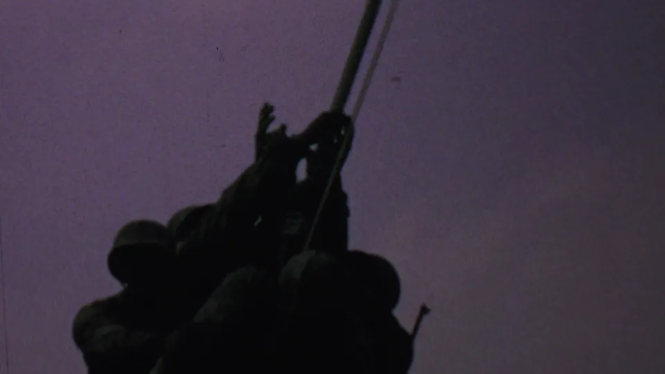 Several silhouetted people appear near the flagpole as the American flag flies above them in the dusk sky, with faces and clothing details not discernible.