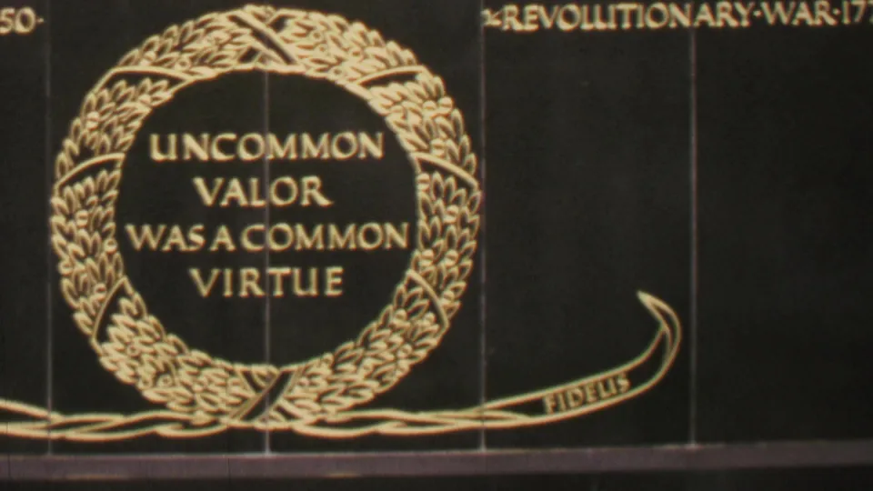 Close view of the Iwo Jima Memorial inscription in Washington, DC, with gold lettering inside a laurel wreath and a partial campaign inscription above.