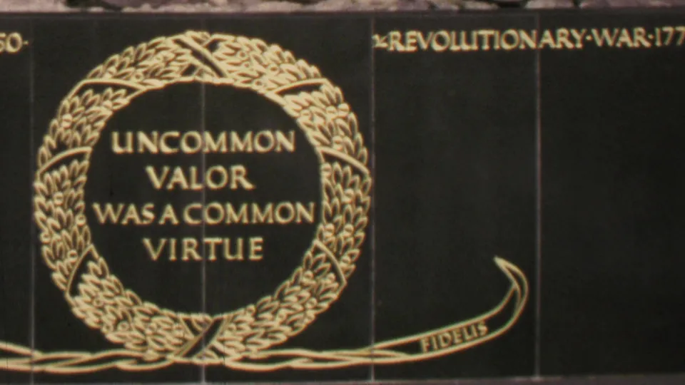 Home-movie close-up of the memorial dedication text and laurel wreath, with partial campaign lettering visible near the upper edge of the frame.