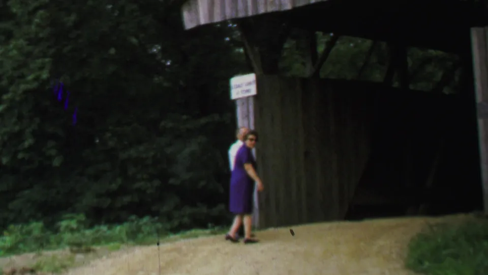1969 United States 8mm frame of an older woman in a purple dress and an older man near the entrance of a rustic wooden covered bridge in a green wooded setting.