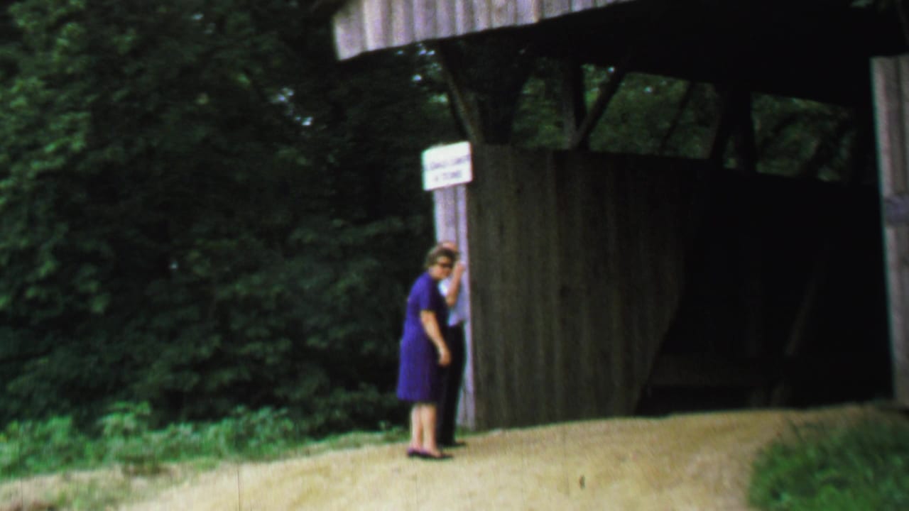 1969 United States 8mm frame showing the woman standing close to a white sign on the wooden support of a covered bridge, with the dark bridge interior behind her.