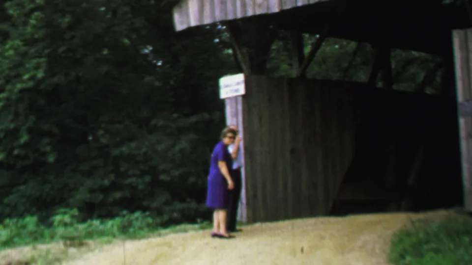 1969 United States 8mm frame showing the woman standing close to a white sign on the wooden support of a covered bridge, with the dark bridge interior behind her.