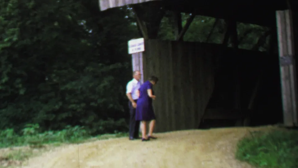 1969 United States 8mm frame of an older man and woman near the wooden entrance post of a covered bridge, appearing to examine or discuss the posted sign.