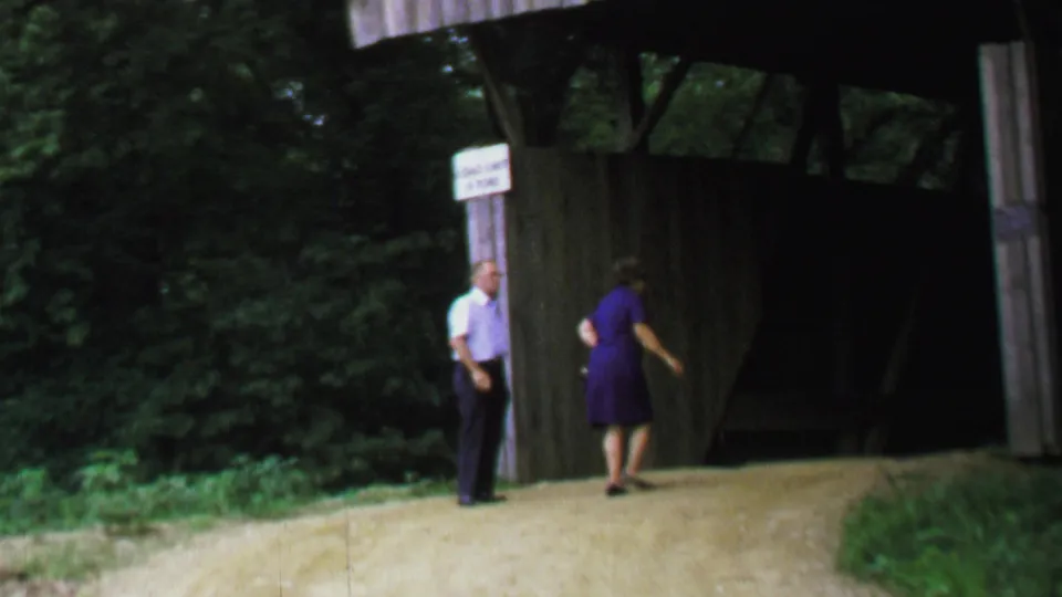 1969 United States 8mm frame showing the woman moving toward the covered bridge opening while the older man remains near the sign and wooded roadside.