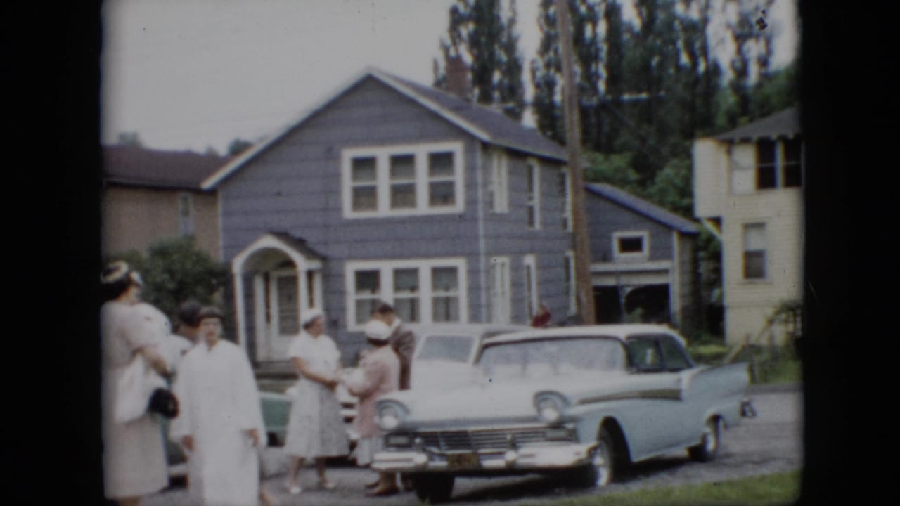 1957 Pennsylvania home-movie frame showing well-dressed adults gathered on a residential street beside a light blue finned sedan and a gray shingled house.