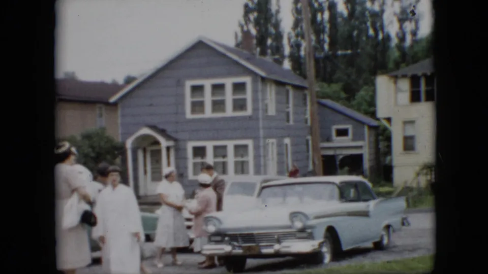 1957 Pennsylvania home-movie frame showing well-dressed adults gathered on a residential street beside a light blue finned sedan and a gray shingled house.