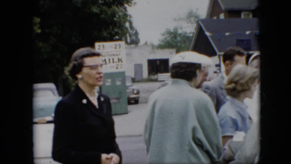 1957 Pennsylvania street scene with a woman in dark formal clothing near other adults, passing cars, and a green milk sign or vending fixture in the background.