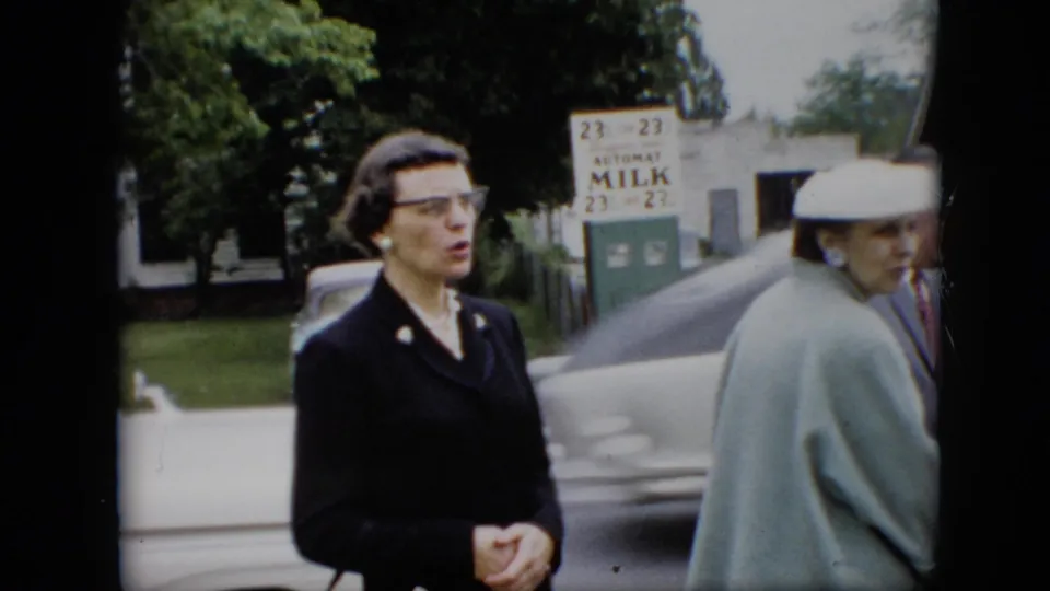 1957 Pennsylvania home-movie frame showing a woman in dark formal clothing on a residential street, with another woman in a pale coat and moving traffic behind her.