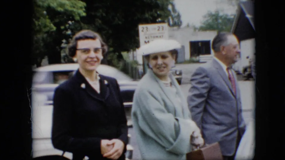 1957 Pennsylvania frame showing two women in formal coats and a man in a suit standing on a residential street, with a green milk sign or vending fixture and blurred passing car behind them.