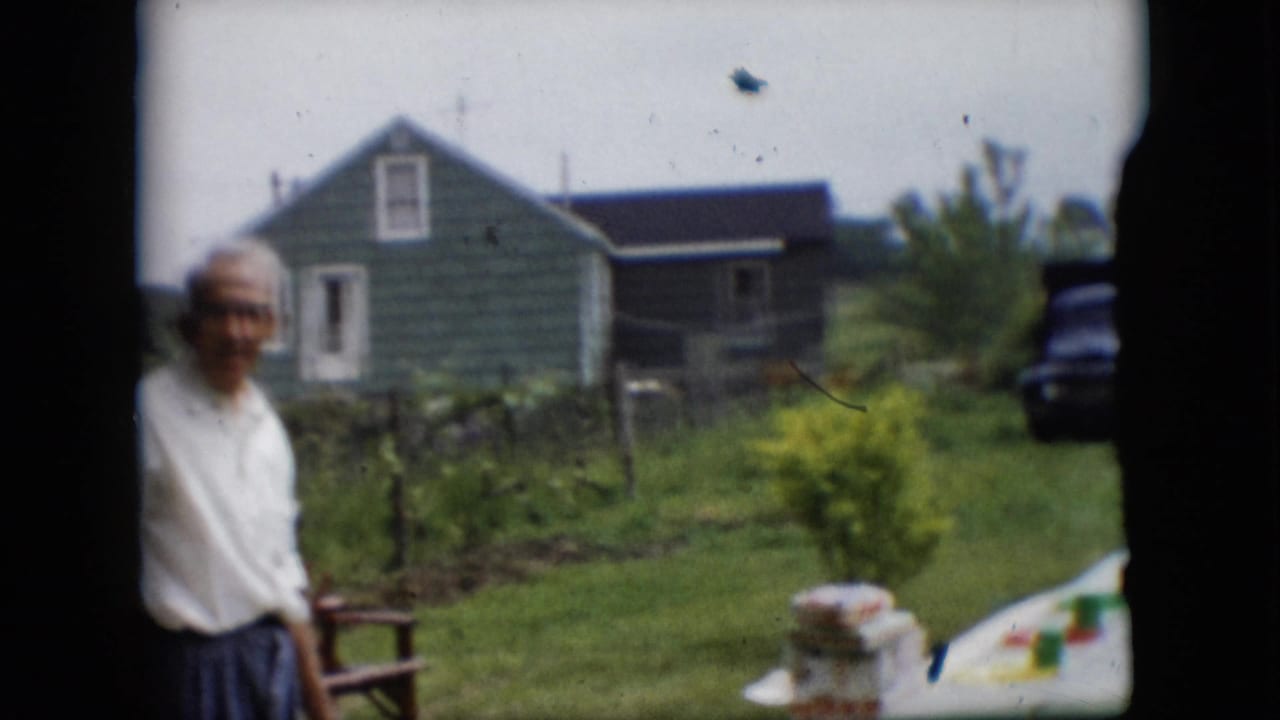 In 1957 Pennsylvania home-movie footage, an older man in a white shirt stands near a white-covered picnic table in a rural yard with a green house, garden area, and dark truck behind him.