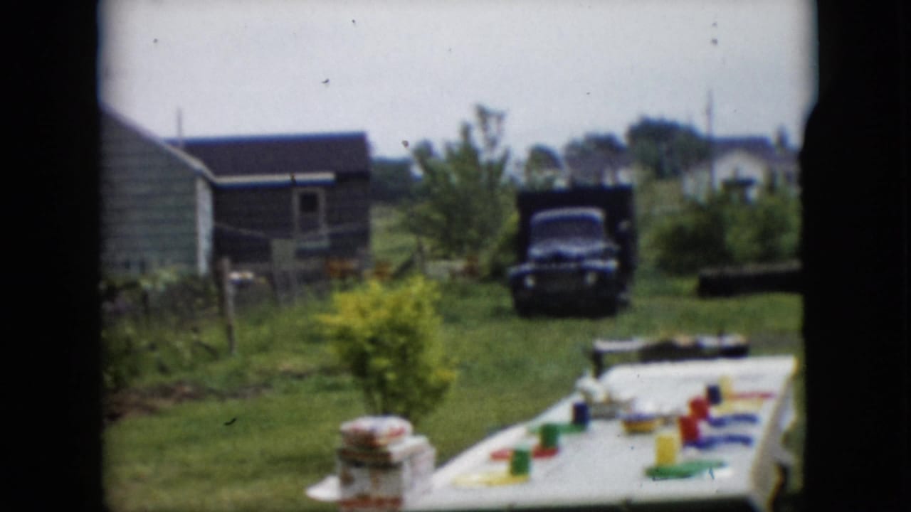 Backyard picnic table with colorful dishes, a green house, lawn, and dark truck in the distance.