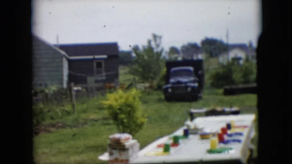 A white-covered outdoor table with colorful cups and plates sits in a rural Pennsylvania backyard, with a green house, garden, lawn, and dark mid-century truck visible beyond it.