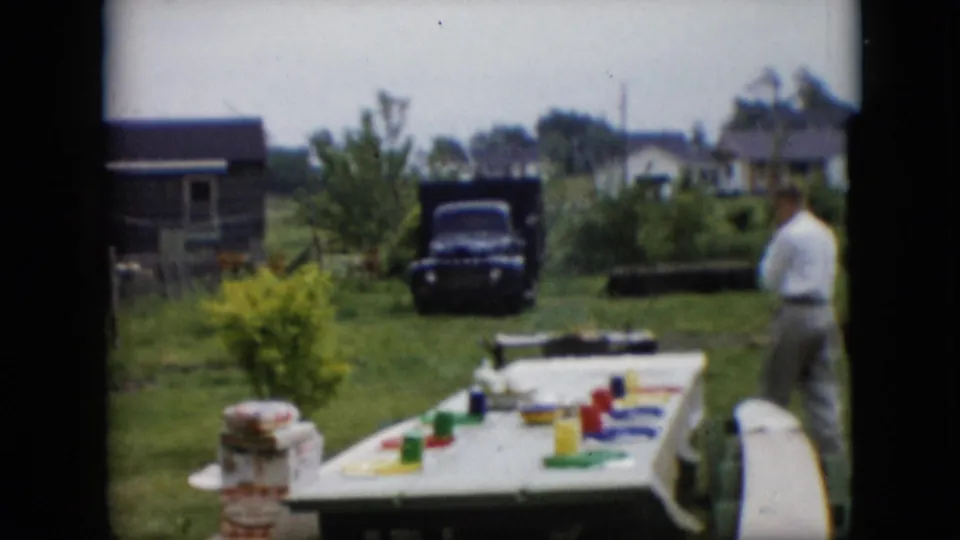 A man in a white shirt and light trousers moves beside a white-covered picnic table in a 1957 Pennsylvania rural yard, with garden rows, houses, and a dark truck in the background.