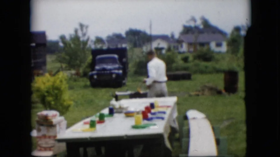 A man viewed from behind stands near a white-covered picnic table in a rural Pennsylvania backyard, with colorful tableware, a dark truck, lawn, shrubs, and neighboring houses visible.