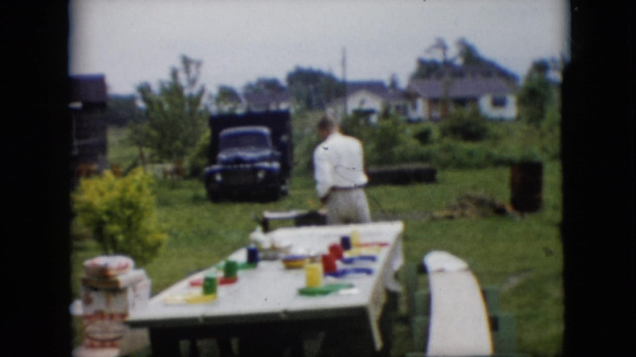 In this 1957 Pennsylvania home-movie frame, an adult man in a white shirt stands near a backyard barbecue area while a picnic table with colorful cups and a dark pickup truck sit in the yard.