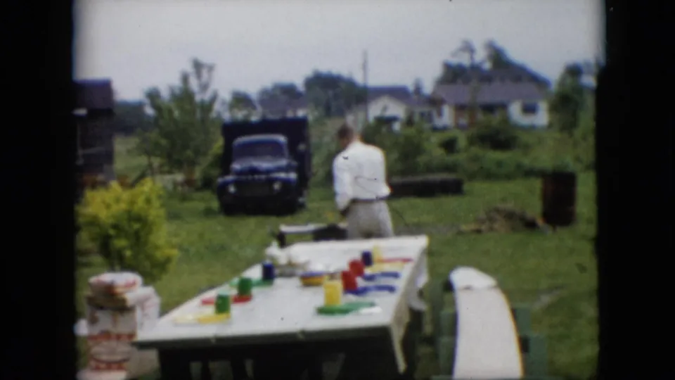 In this 1957 Pennsylvania home-movie frame, an adult man in a white shirt stands near a backyard barbecue area while a picnic table with colorful cups and a dark pickup truck sit in the yard.