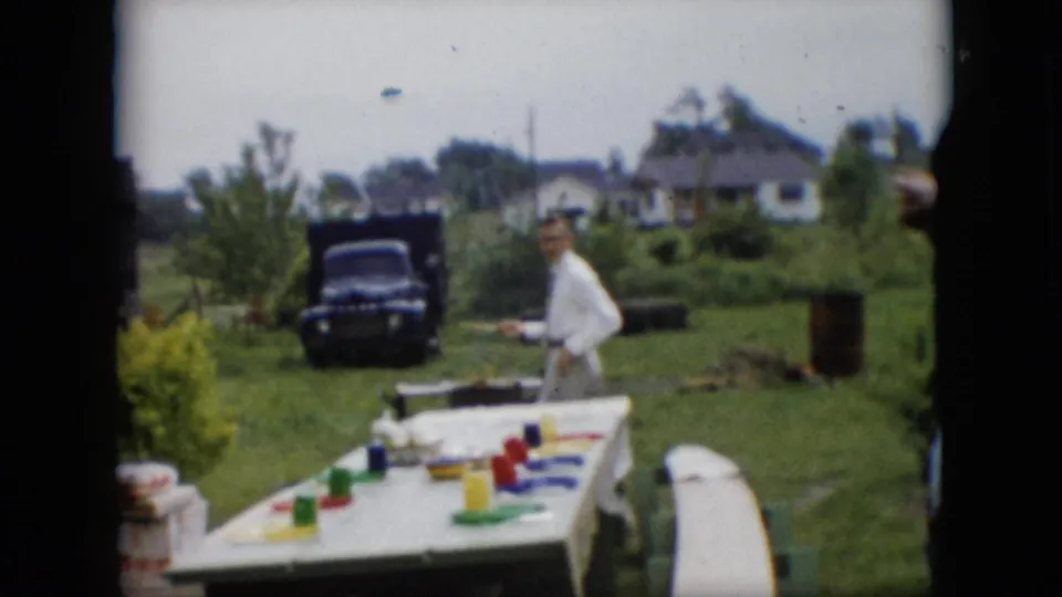 An adult man in a white shirt stands beside the grill area in a Pennsylvania backyard, with colorful cups on a white-covered picnic table and a dark pickup truck in the background.