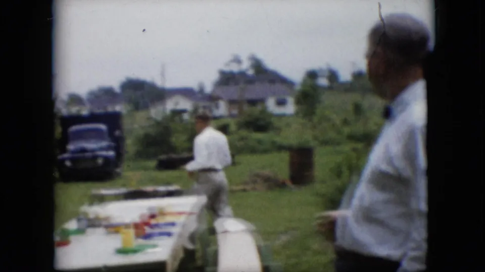Two adult men in white shirts appear in the backyard cookout scene, one near the grill and another closer to the camera, with the picnic table, pickup truck, grass, and houses visible around them.