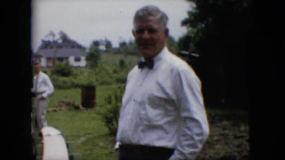 An adult man wearing a white shirt and bow tie stands close to the camera in a 1957 Pennsylvania backyard, with another man, a grill area, lawn, and distant houses behind him.