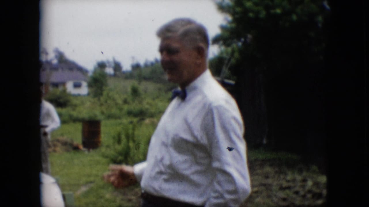 In metadata-supported 1957 Pennsylvania footage, a man in a white dress shirt and black bow tie stands outdoors on a lawn with houses and greenery behind him.