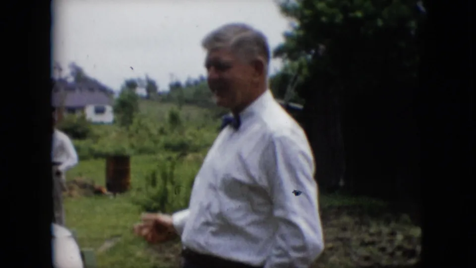 In metadata-supported 1957 Pennsylvania footage, a man in a white dress shirt and black bow tie stands outdoors on a lawn with houses and greenery behind him.