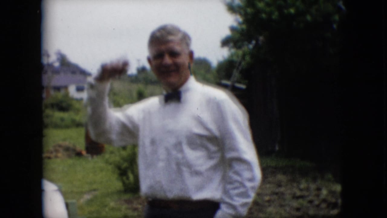 Smiling man in a white shirt and black bow tie waving toward the camera outdoors.