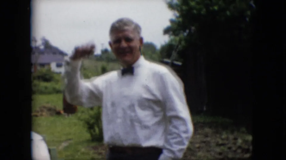 A smiling man in a white dress shirt and black bow tie gestures toward the camera in an outdoor home-movie scene from metadata-supported 1957 Pennsylvania.
