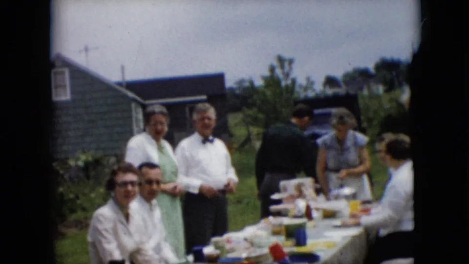 Several adults in 1950s dress gather around a long table with food and drinks on a grassy lawn in metadata-supported Pennsylvania home-movie footage.