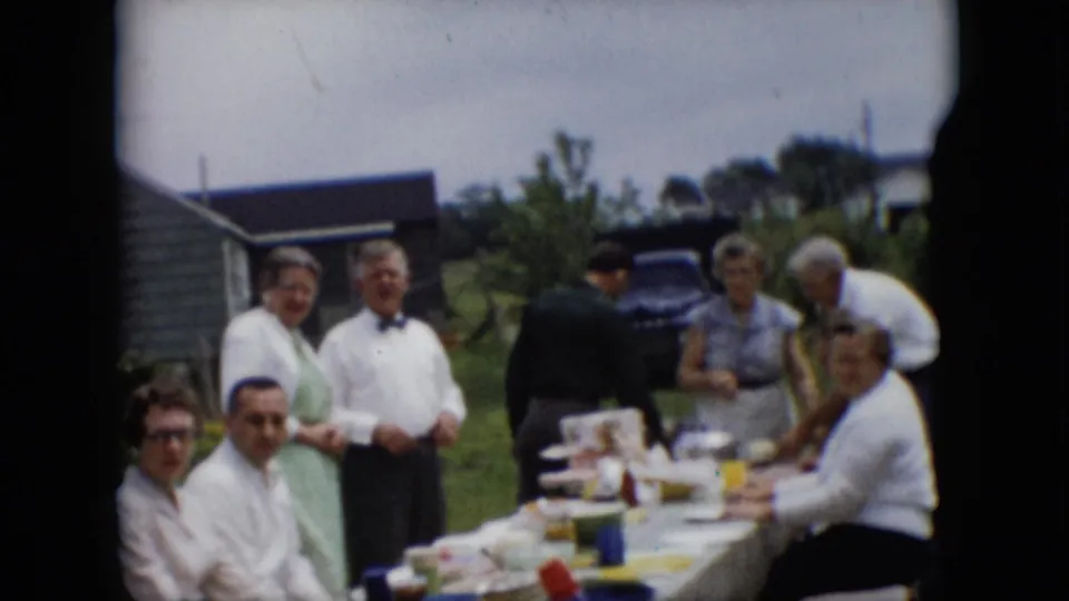 Adults stand and sit around a table of food outdoors, with some looking toward the camera during a metadata-supported 1957 Pennsylvania gathering.