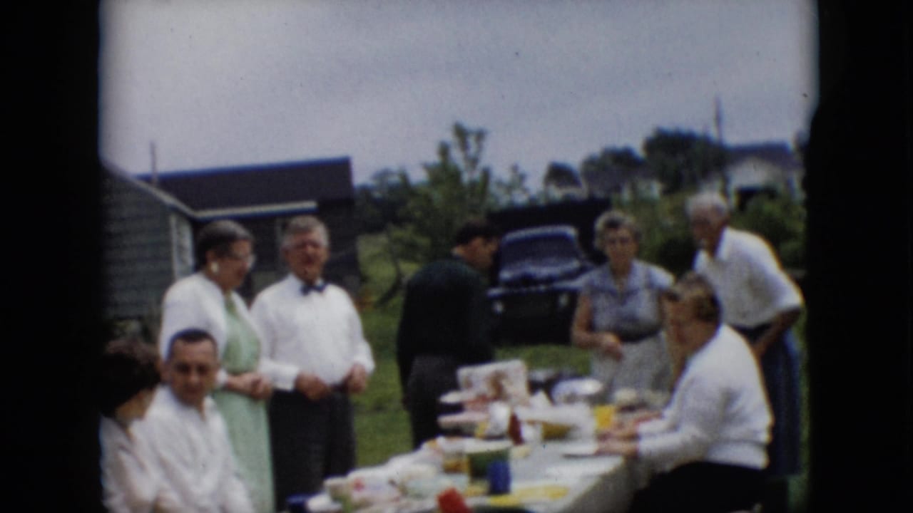 1957 Pennsylvania 8mm home-movie frame showing adults in mid-century clothing standing and sitting around a picnic table with food in a grassy yard.