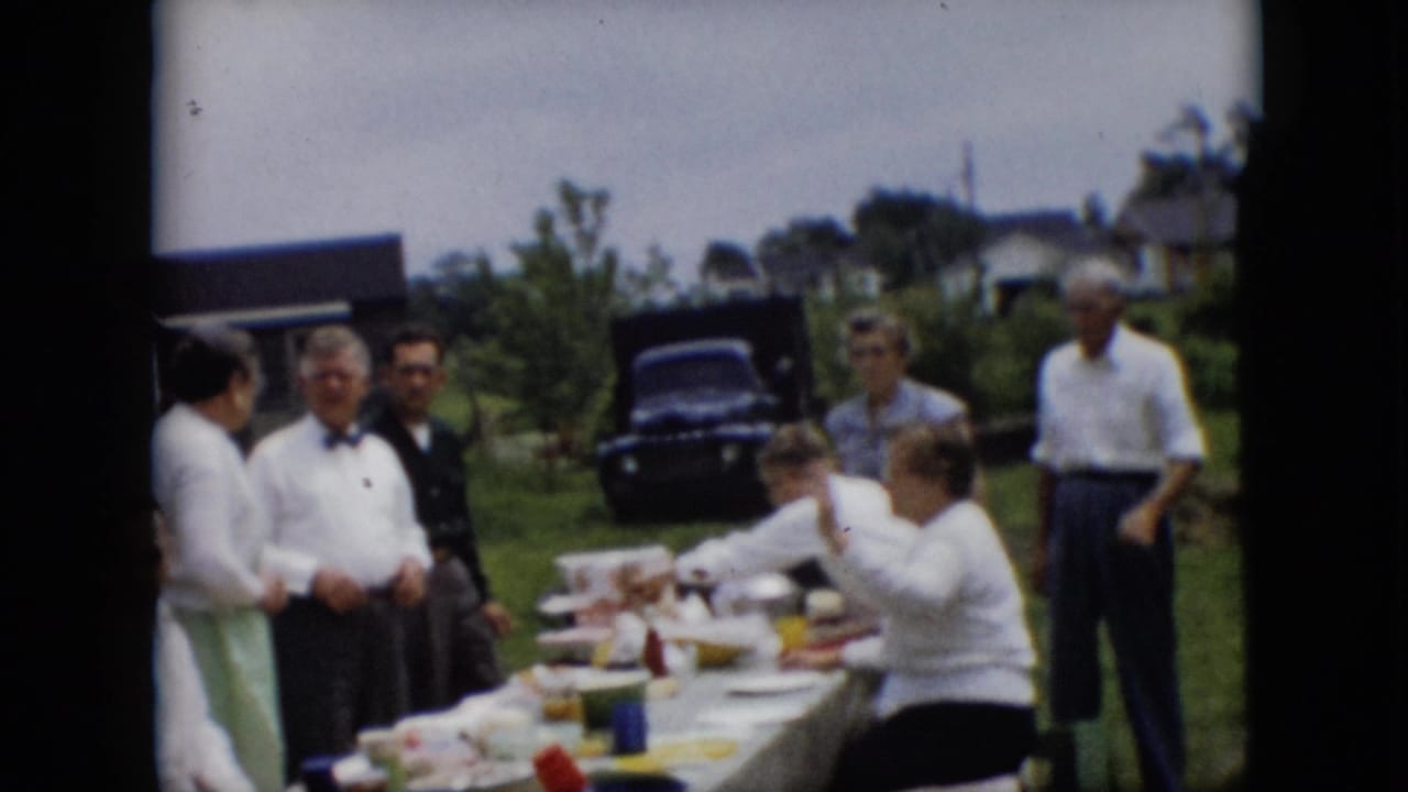 Several adults stand and sit beside a picnic table with food outdoors.