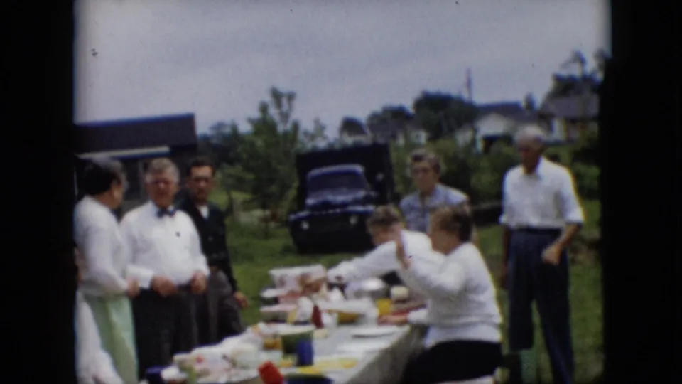 Adults in 1950s attire stand and sit near a long outdoor picnic table, with a dark car and houses visible behind them in the Pennsylvania home-movie setting.