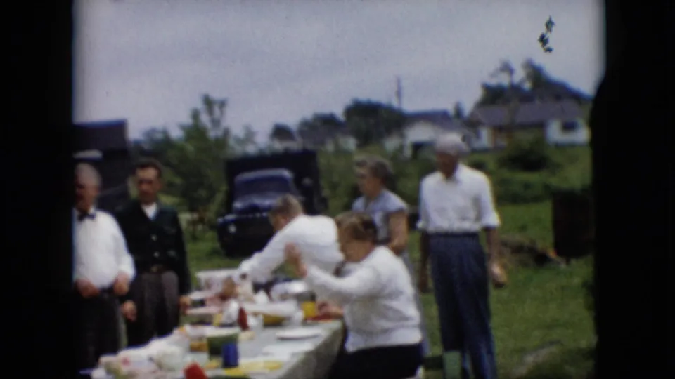 A 1957 Pennsylvania backyard picnic scene shows adults serving food and talking around a long table, with a dark mid-century car and homes in the background.